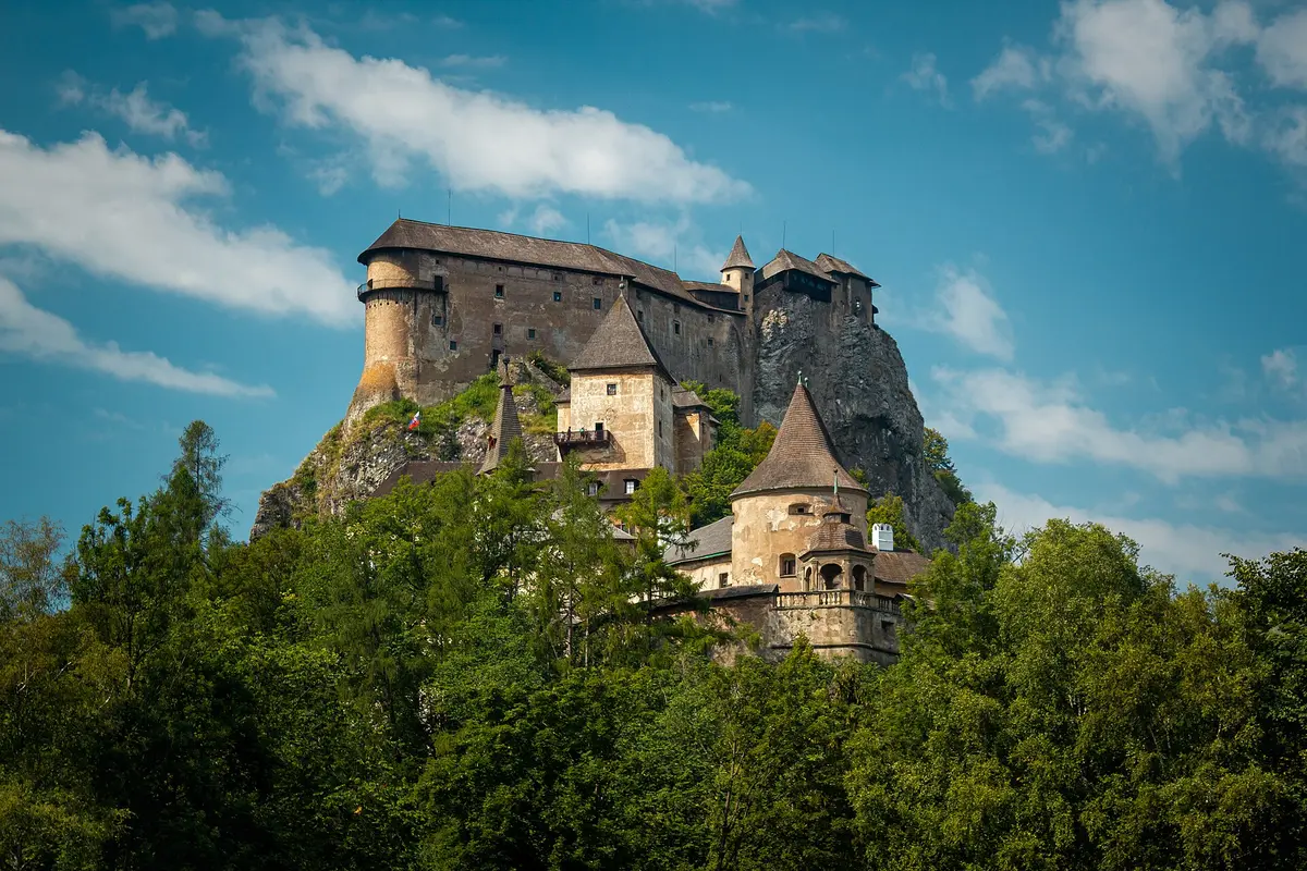 Orava Castle's towers and palace wings rising up a bare limestone cliff above a band of green forest, under a blue sky