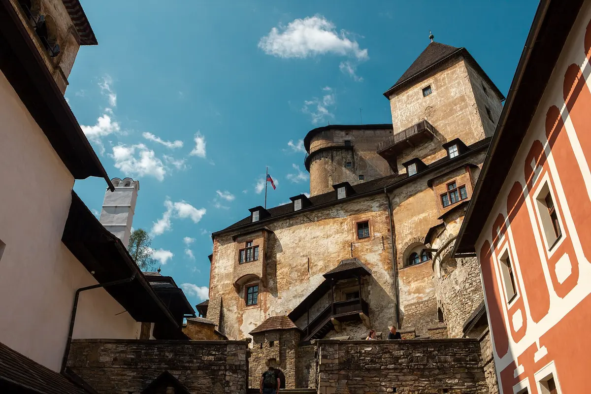 Looking up from a stone-walled courtyard inside Orava Castle at a tall tower and older wings, with a red and white painted building on the right